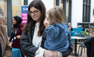 A young mother with dark hair and glasses holds a little toddler in her arms